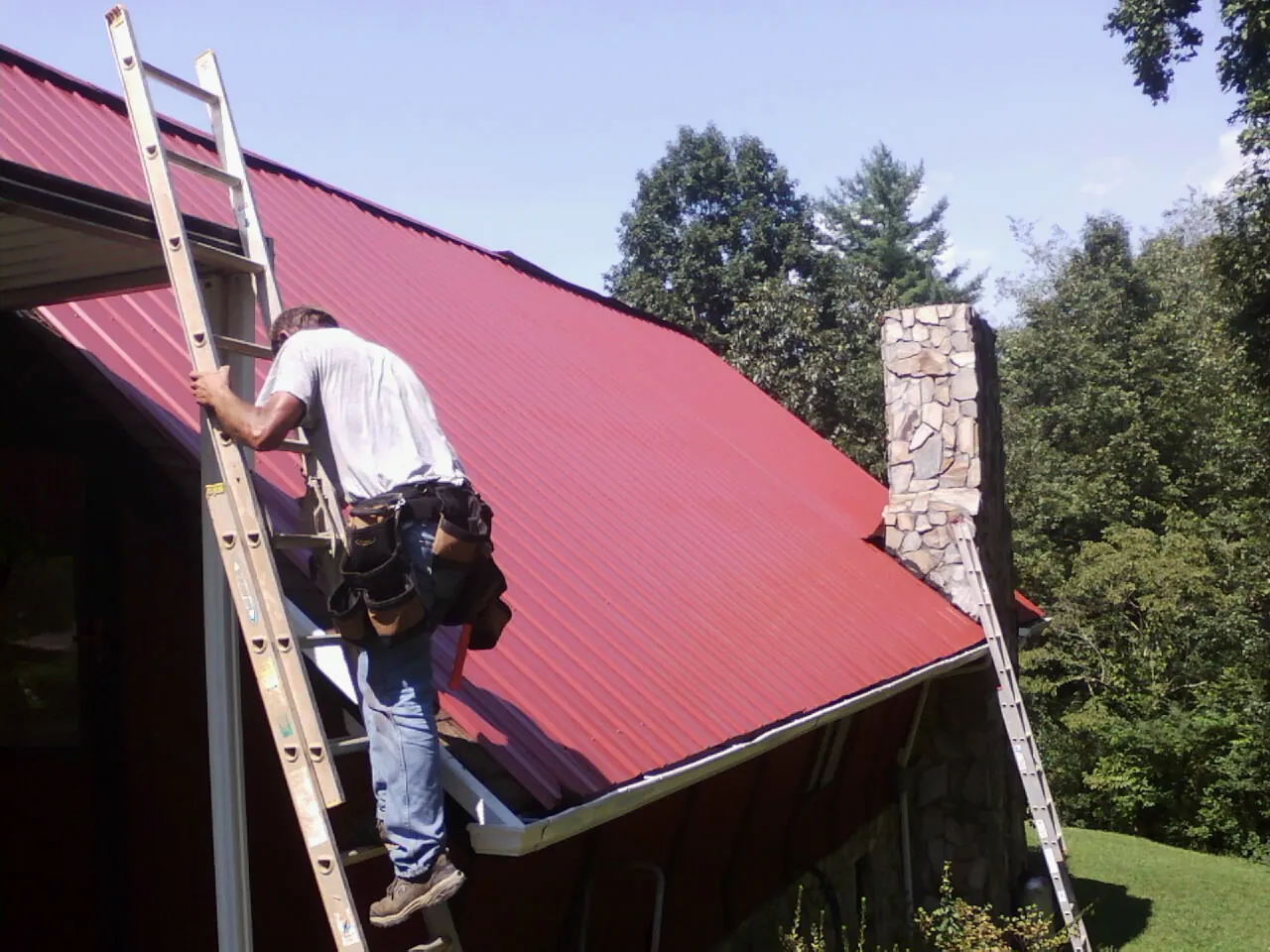 Roofer on a ladder beside a red metal roof and stone chimney — Soffit & Fascia Repair by Roofing Brothers in Beech Island